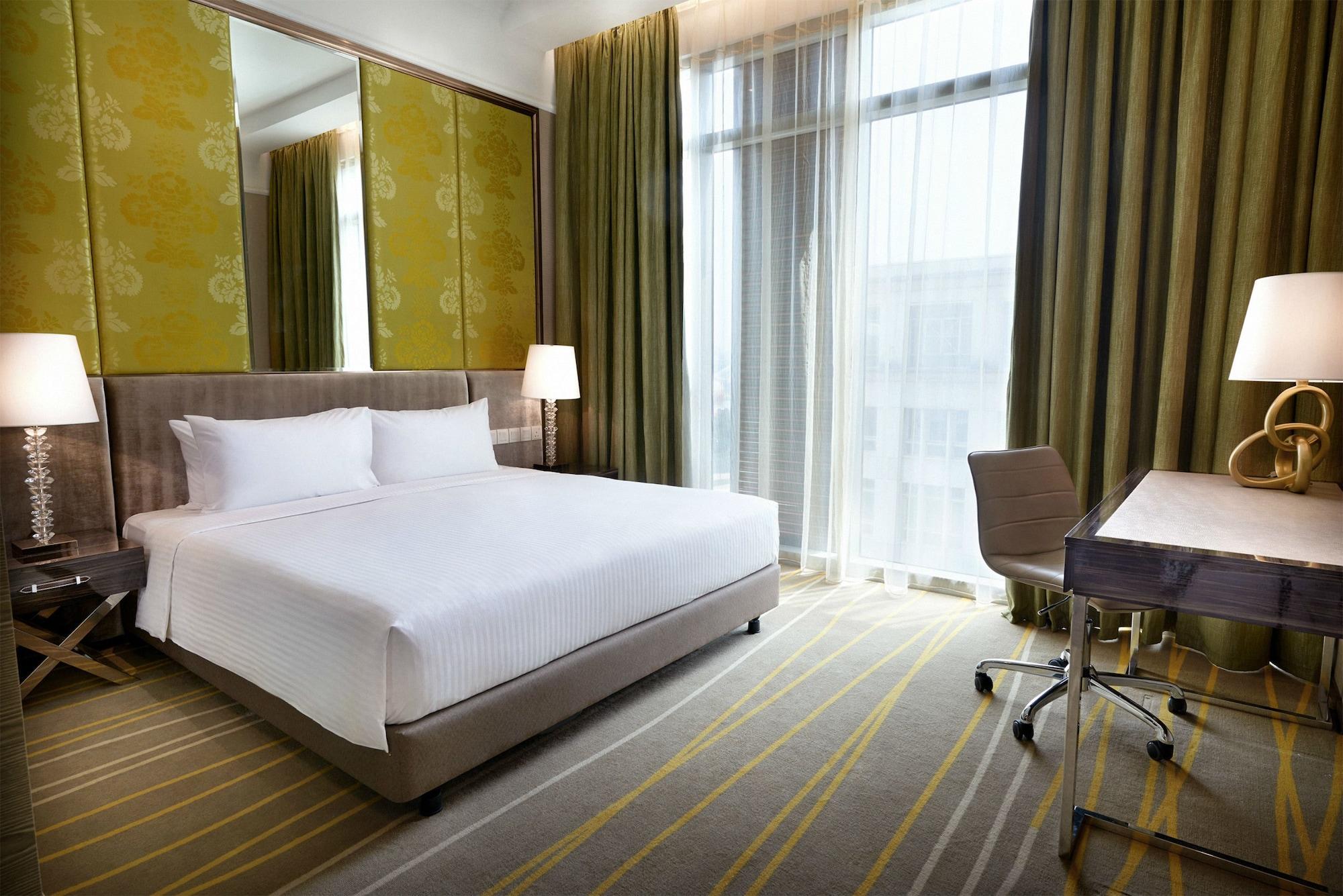 Hotel Dorsett Putrajaya Exteriér fotografie The photo shows a modern hotel room. It features a large bed with white bedding positioned against a wall. The room has tall windows covered with sheer curtains, letting in natural light. There are two bedside lamps on either side of the bed. A desk 