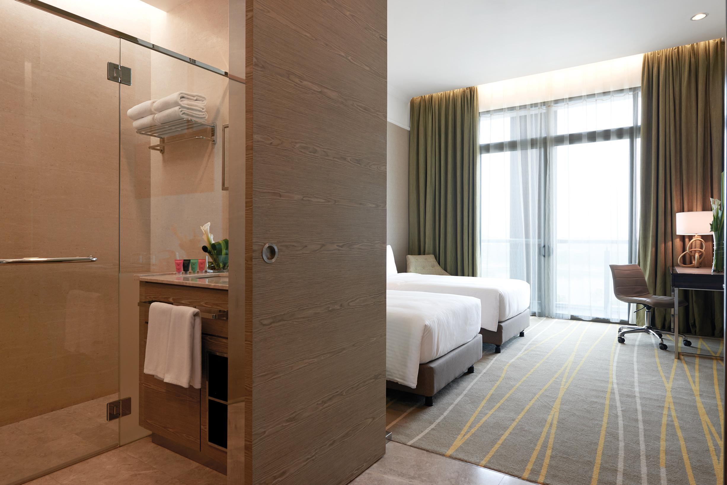 Hotel Dorsett Putrajaya Exteriér fotografie The photo shows a modern hotel room with a clean and inviting design. On the left side, there is a bathroom area featuring a glass shower and some neatly arranged towels. The bathroom has a sink with toiletries and a small shelf. 

On the right side,