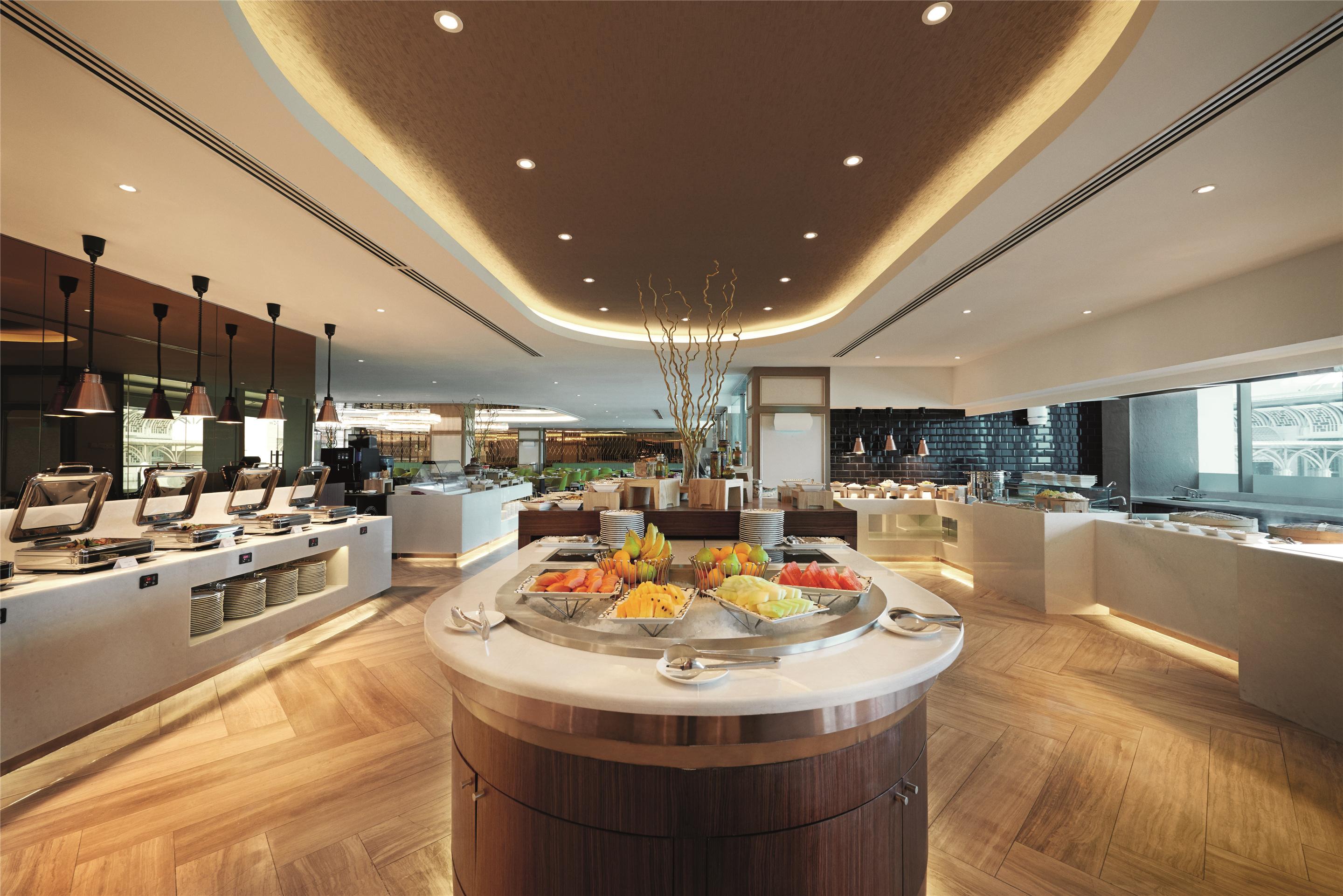 Hotel Dorsett Putrajaya Exteriér fotografie The photo shows a modern buffet restaurant interior. In the center, there's a circular display with a variety of fresh fruits arranged attractively. Surrounding this are several serving stations and food displays that feature a clean, contemporary de