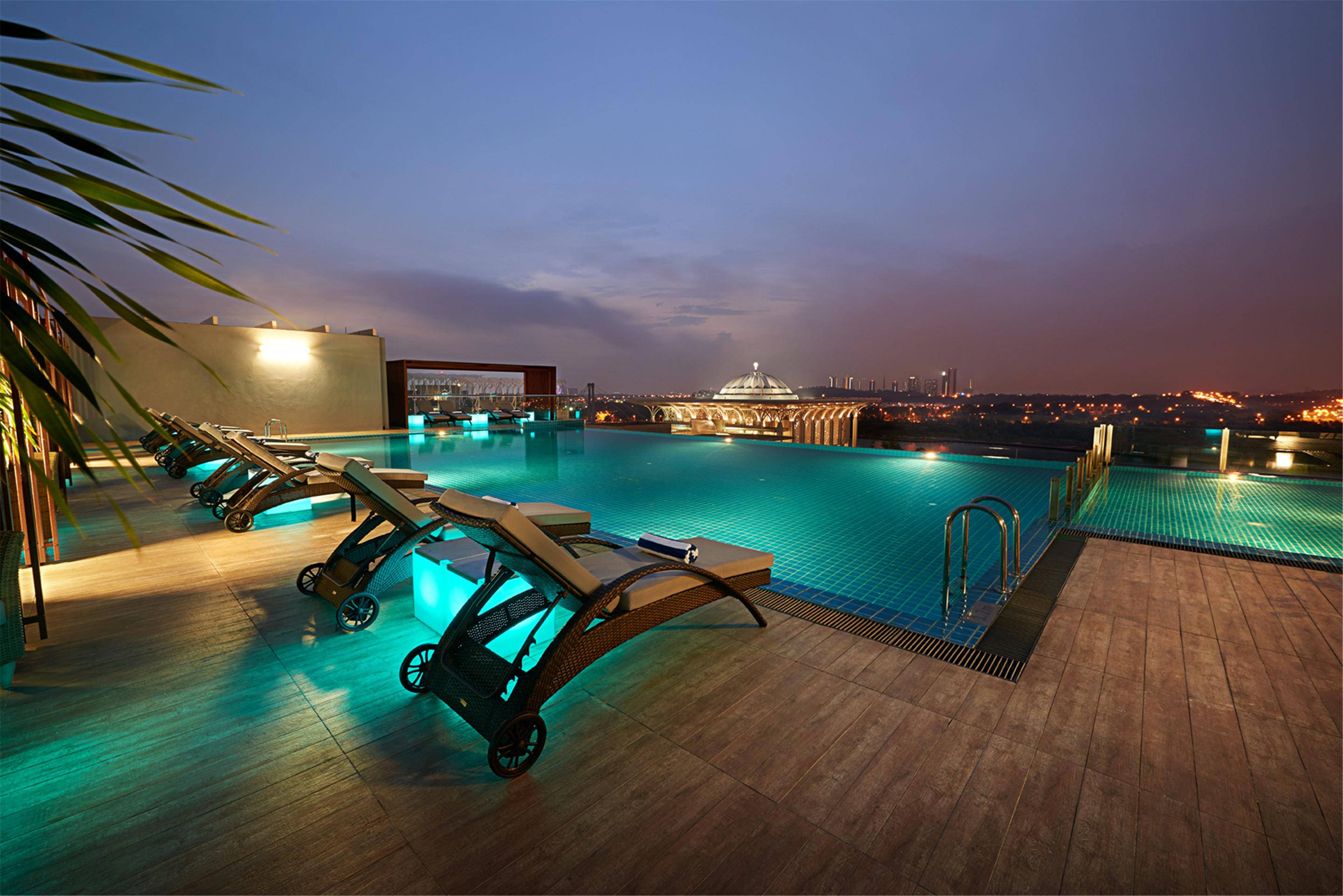 Hotel Dorsett Putrajaya Exteriér fotografie The photo shows a rooftop pool area during twilight, featuring a serene and modern design. There are several lounge chairs positioned around the pool, which is illuminated with soft blue lights, creating a relaxing atmosphere. In the background, city