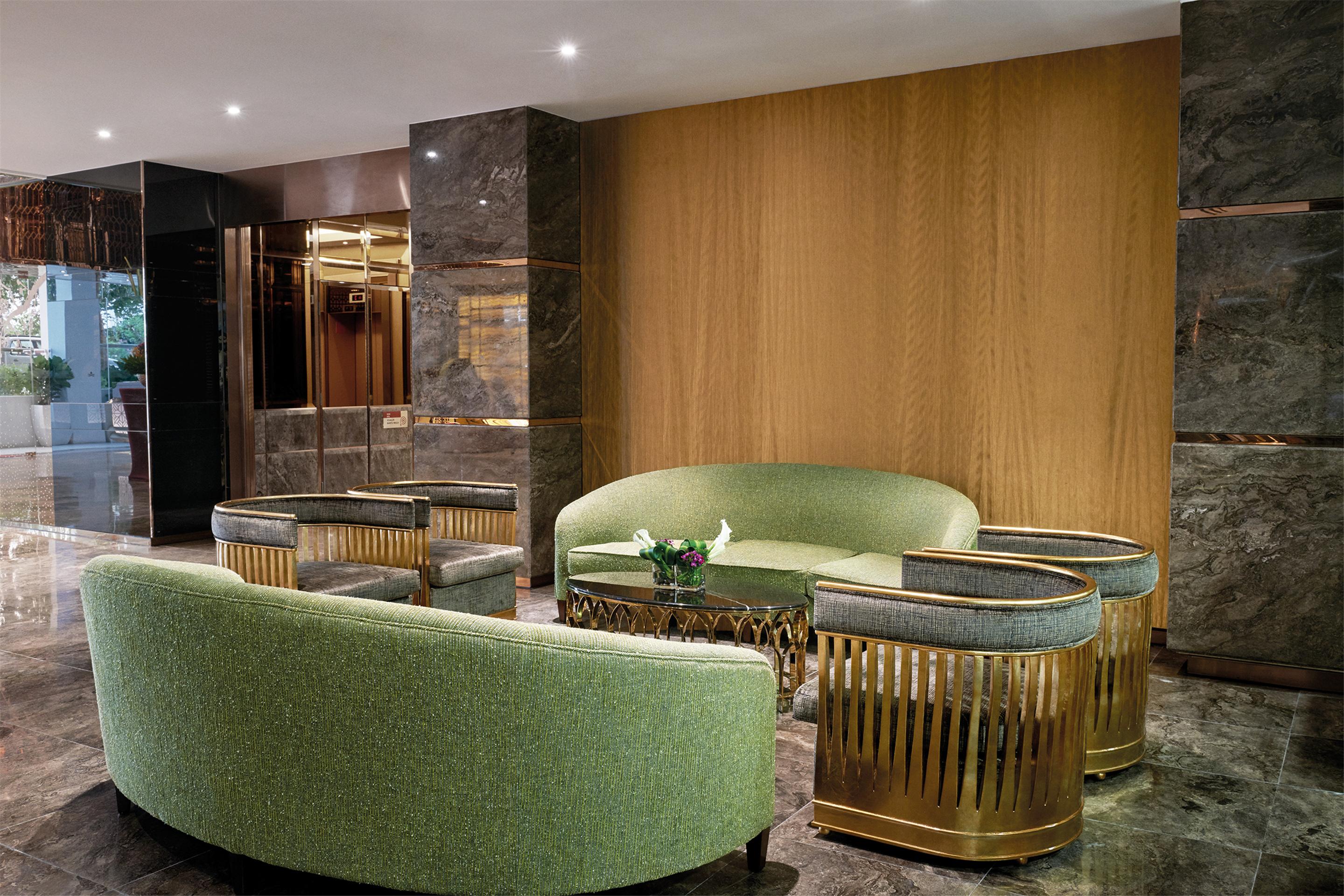 Hotel Dorsett Putrajaya Exteriér fotografie The photo shows a stylish lounge area with modern furniture. There is a large, curved green sofa accompanied by matching green accent chairs. The seating is arranged around a unique gold and glass coffee table. The backdrop features a wooden wall wit