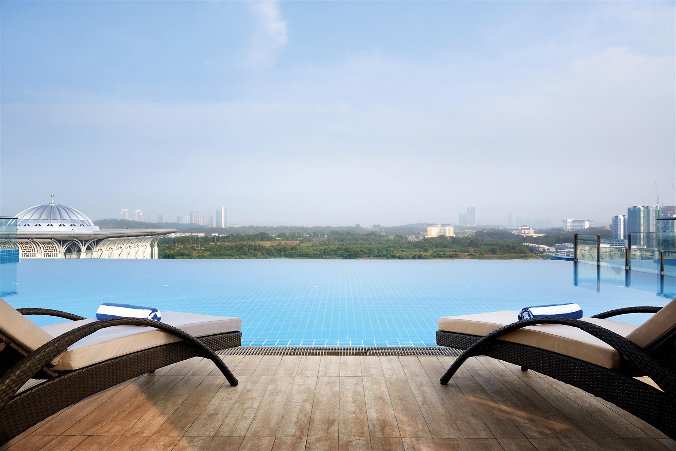 Hotel Dorsett Putrajaya Exteriér fotografie The photo depicts a luxurious outdoor space featuring an infinity pool that extends toward the horizon, blending seamlessly with the sky. In the foreground, there are two sun loungers with rolled towels on each, positioned on a wooden deck. The backd