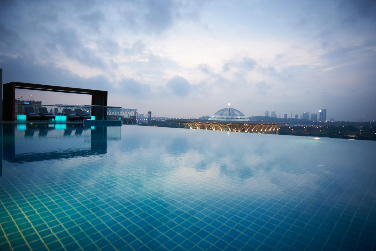 Hotel Dorsett Putrajaya Exteriér fotografie The photo shows a serene infinity pool with a calm, reflective surface, blending seamlessly with the sky. In the background, there's a view of a cityscape with buildings and a large dome structure, possibly a stadium or an arena. The atmosphere appea