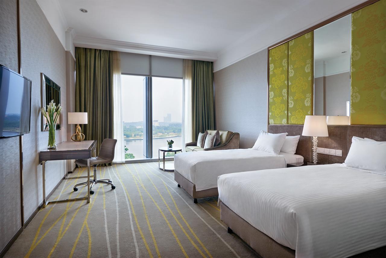 Hotel Dorsett Putrajaya Pokoj fotografie The photo shows a modern hotel room featuring two single beds with white linens. There is a stylish desk and chair positioned beside a large window that allows natural light to fill the space. The window is adorned with long, light green curtains. A 