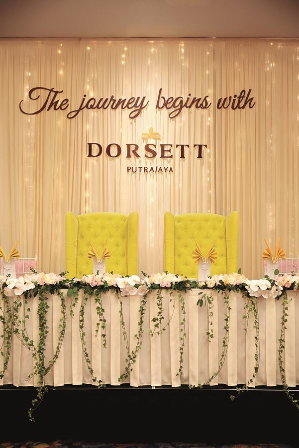 Hotel Dorsett Putrajaya Exteriér fotografie The photo features a decorative setup, likely for an event. In the center, there are two prominent yellow chairs with tufted upholstery, positioned against a backdrop of soft draping. Above the chairs, the name "DORSETT PUTRAJAYA" is displayed, indic