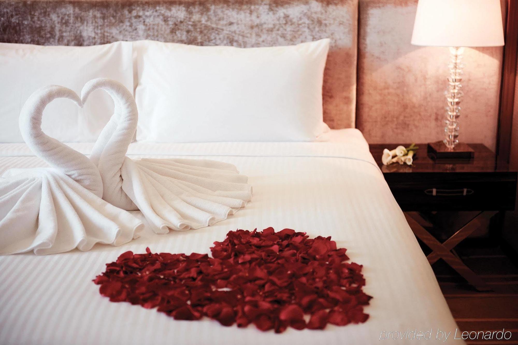 Hotel Dorsett Putrajaya Exteriér fotografie The photo displays a romantic hotel bed setting. On the neatly arranged white sheets, two swan-shaped towel sculptures are positioned, resembling a heart. Surrounding the towels, there is a heart shape made of red rose petals. To the side, there is a