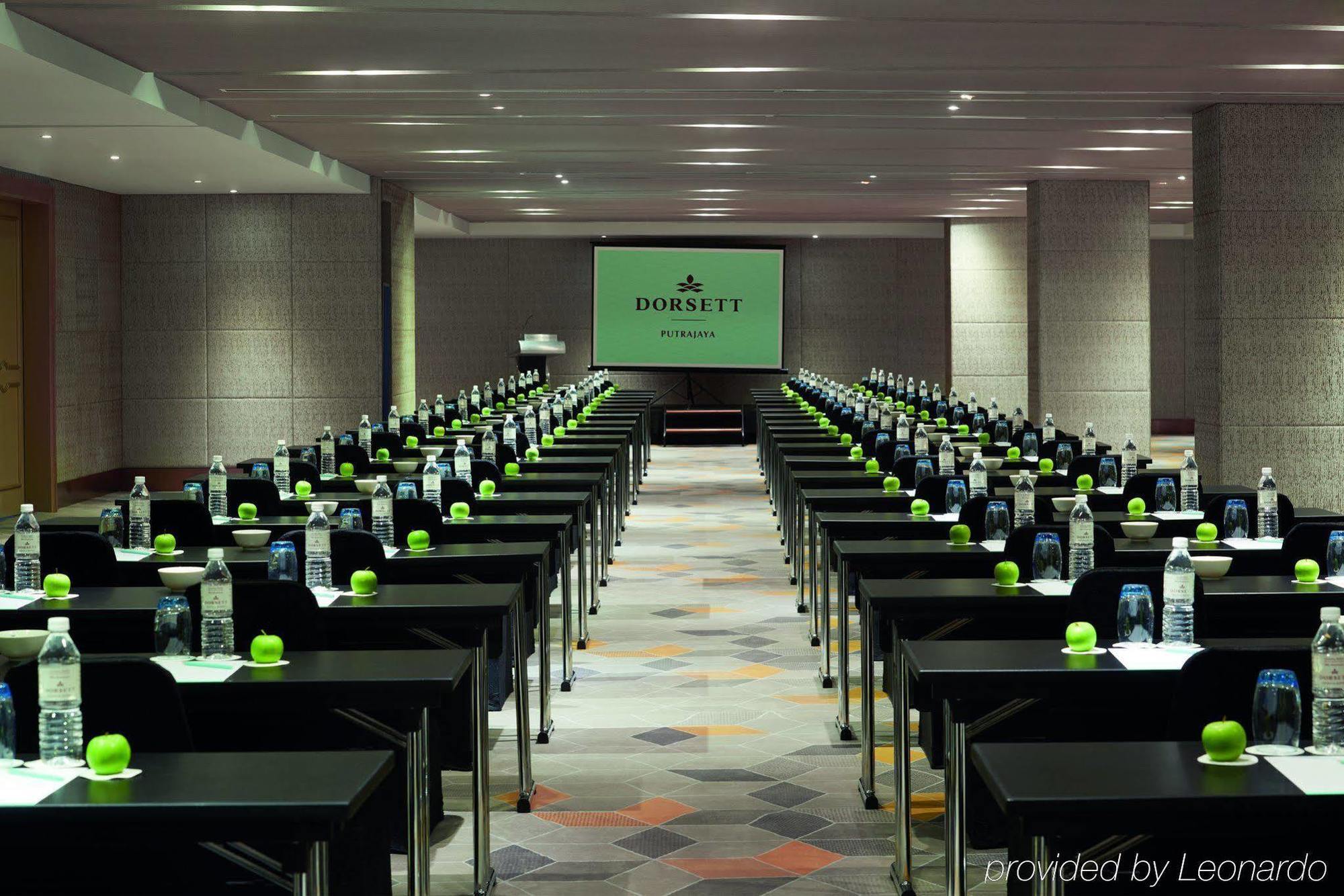 Hotel Dorsett Putrajaya Exteriér fotografie The photo shows a conference room set up for a meeting or event. There are multiple rows of black tables arranged neatly, each equipped with a green object, possibly a plant or decorative item, and a bottle of water. In the center of the room, there 