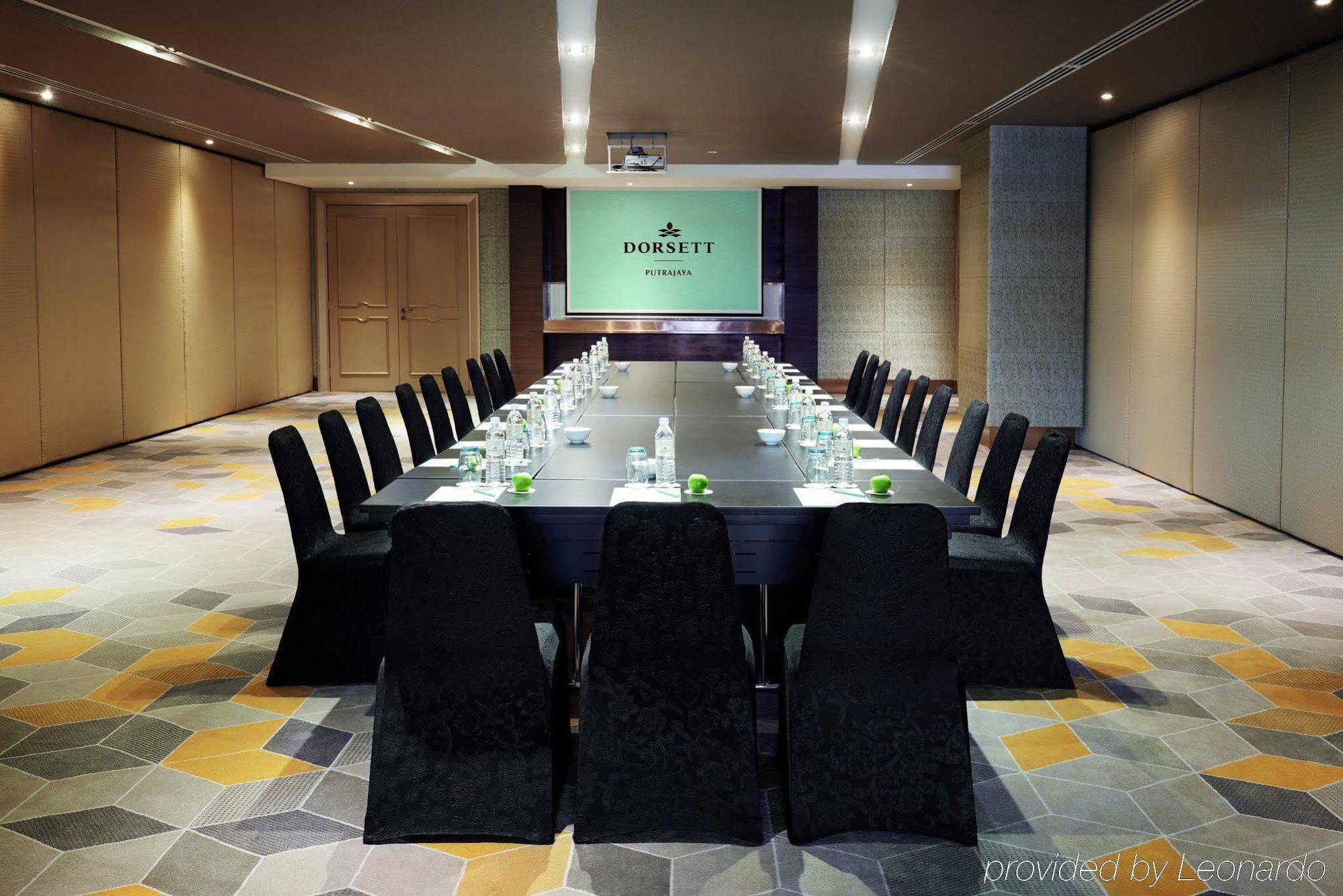 Hotel Dorsett Putrajaya Exteriér fotografie The photo shows a modern conference room set up for a meeting. A long, dark wooden table is surrounded by black chairs with a sleek design. On the table, there are bottles of water and small green plants or decorative items. At the head of the room, 