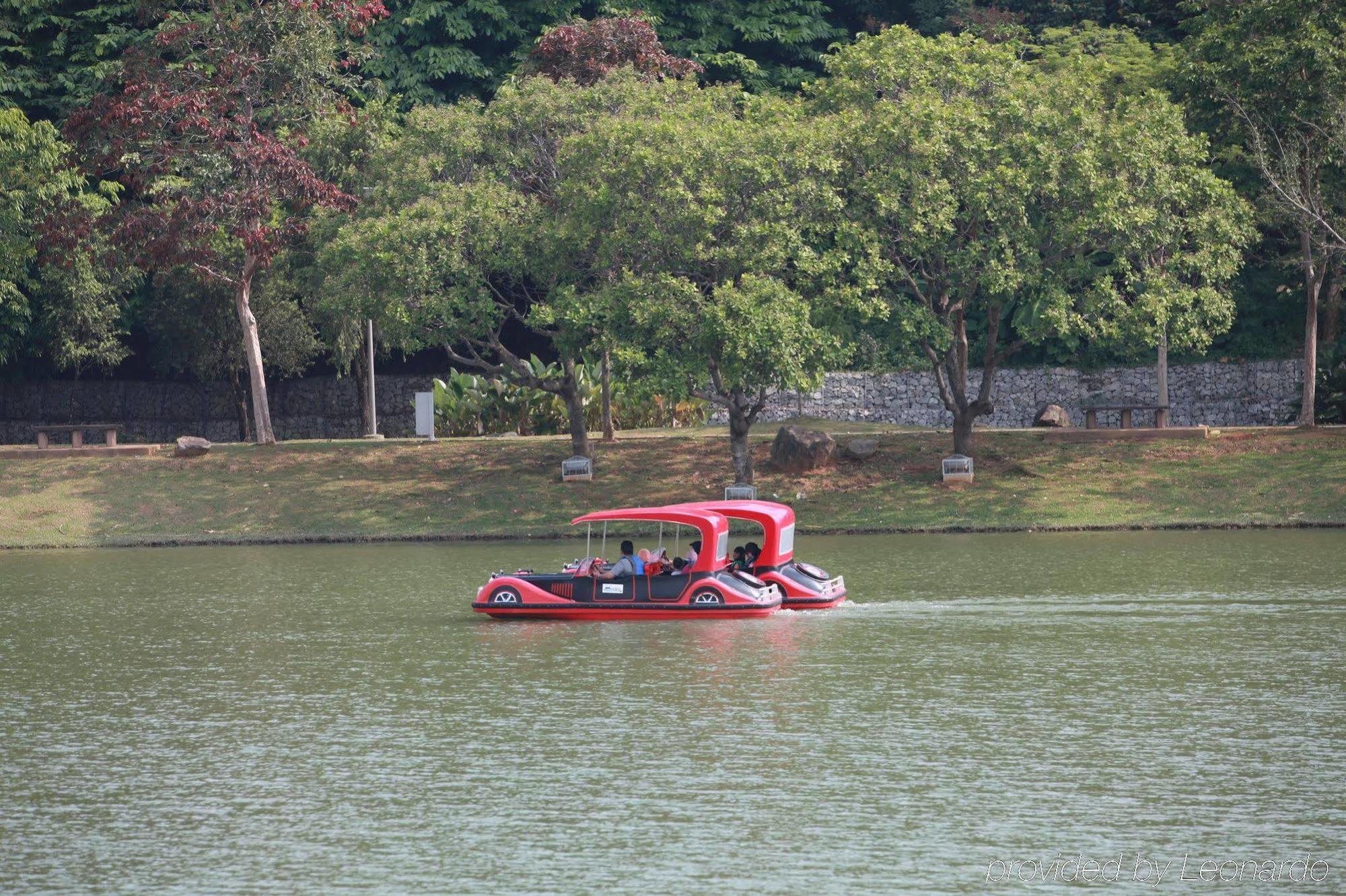 Hotel Dorsett Putrajaya Exteriér fotografie The photo shows a red boat gliding across a calm lake. The boat appears to be a pedal or leisure boat, with several people on board. In the background, there are trees and greenery, contributing to a serene natural setting. The water reflects the sur