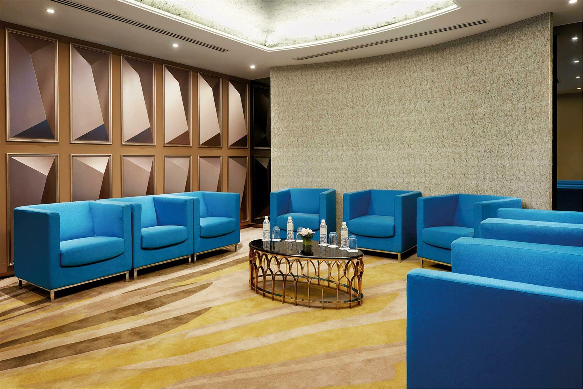 Hotel Dorsett Putrajaya Exteriér fotografie The photo shows a modern meeting or lounge area. It features bright blue chairs arranged around a central table. The table has decorative elements and bottles of water on it. The room is designed with stylish walls that have a geometric pattern and a