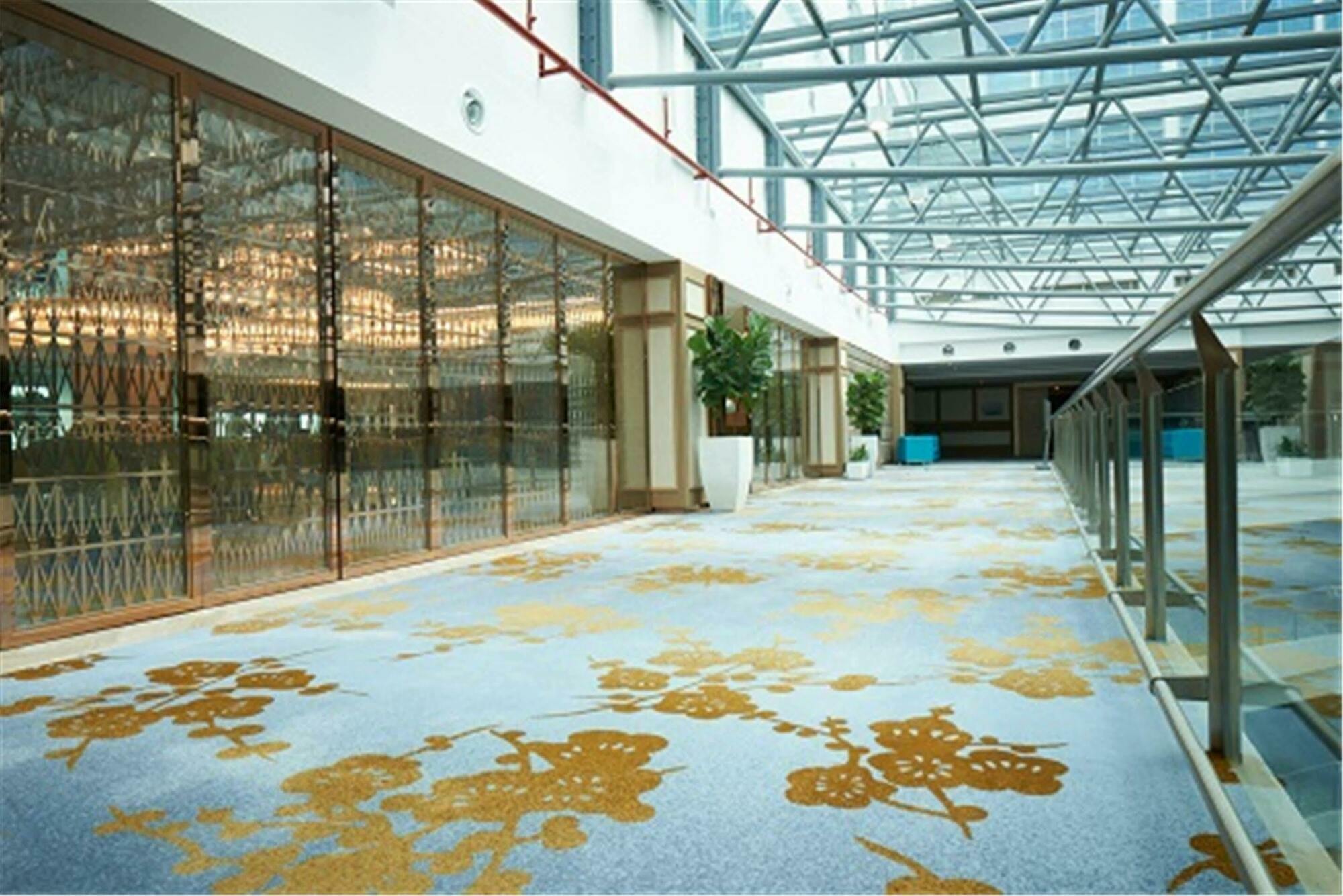 Hotel Dorsett Putrajaya Exteriér fotografie The photo shows a spacious, brightly lit interior setting, likely a corridor or lobby area. The ceiling is made of glass, allowing natural light to flood the space. The flooring features a decorative carpet with floral patterns in shades of gold. To 