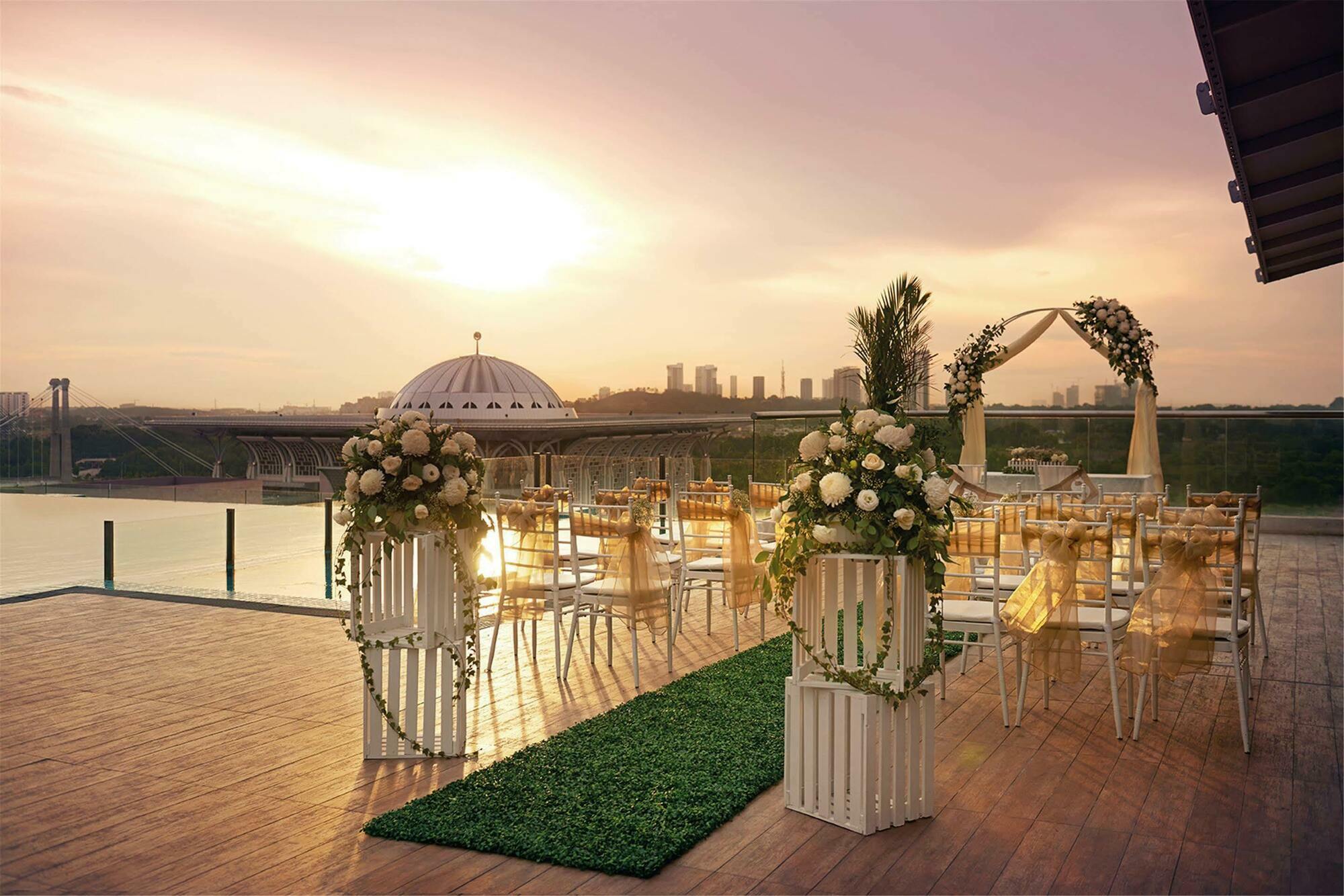 Hotel Dorsett Putrajaya Exteriér fotografie The photo shows a beautifully designed outdoor wedding venue. In the foreground, there are elegant arrangements of flowers on white pedestals, creating a romantic atmosphere. There is a pathway lined with green grass leading to an arch adorned with f