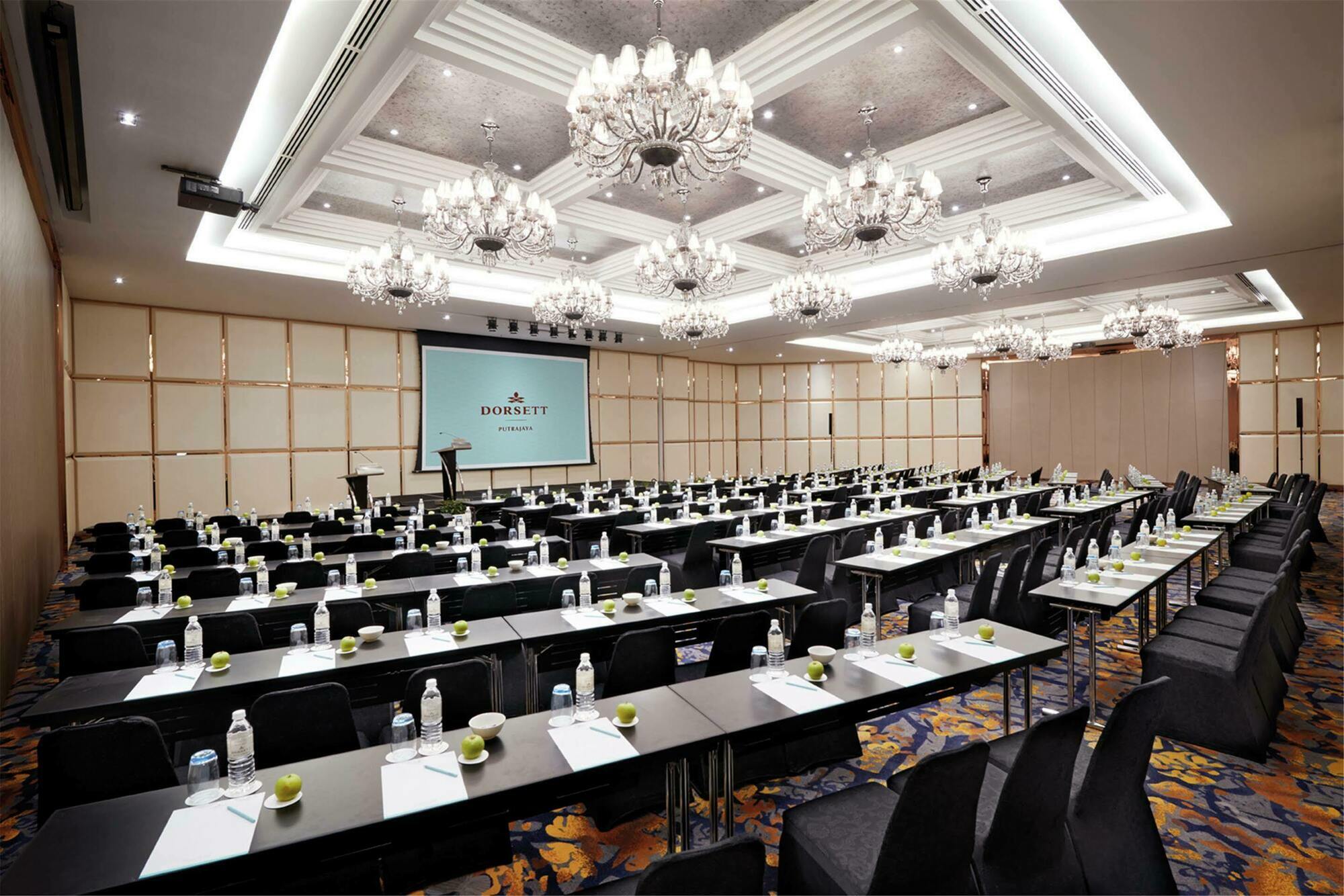 Hotel Dorsett Putrajaya Exteriér fotografie The photo shows a large conference room set up for an event. It features a number of black tables arranged in rows, each equipped with water bottles and small green apples. The room has a modern and elegant decor, highlighted by ornate chandeliers ha